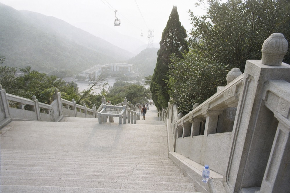 An exposed stone stairway climbing toward the Buddha under a hazy summer sky on Lantau, no text, no logos, no people