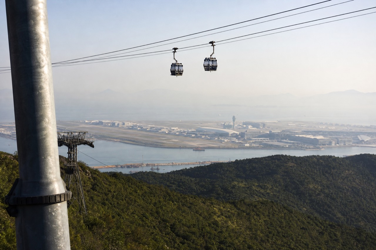 Cable car cabins crossing green Lantau hillside above the airport island, no text, no logos, no people.