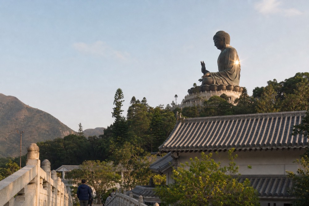 Bronze Tian Tan Buddha statue seen from below with the monastery roofs in the foreground, no text, no logos, no people