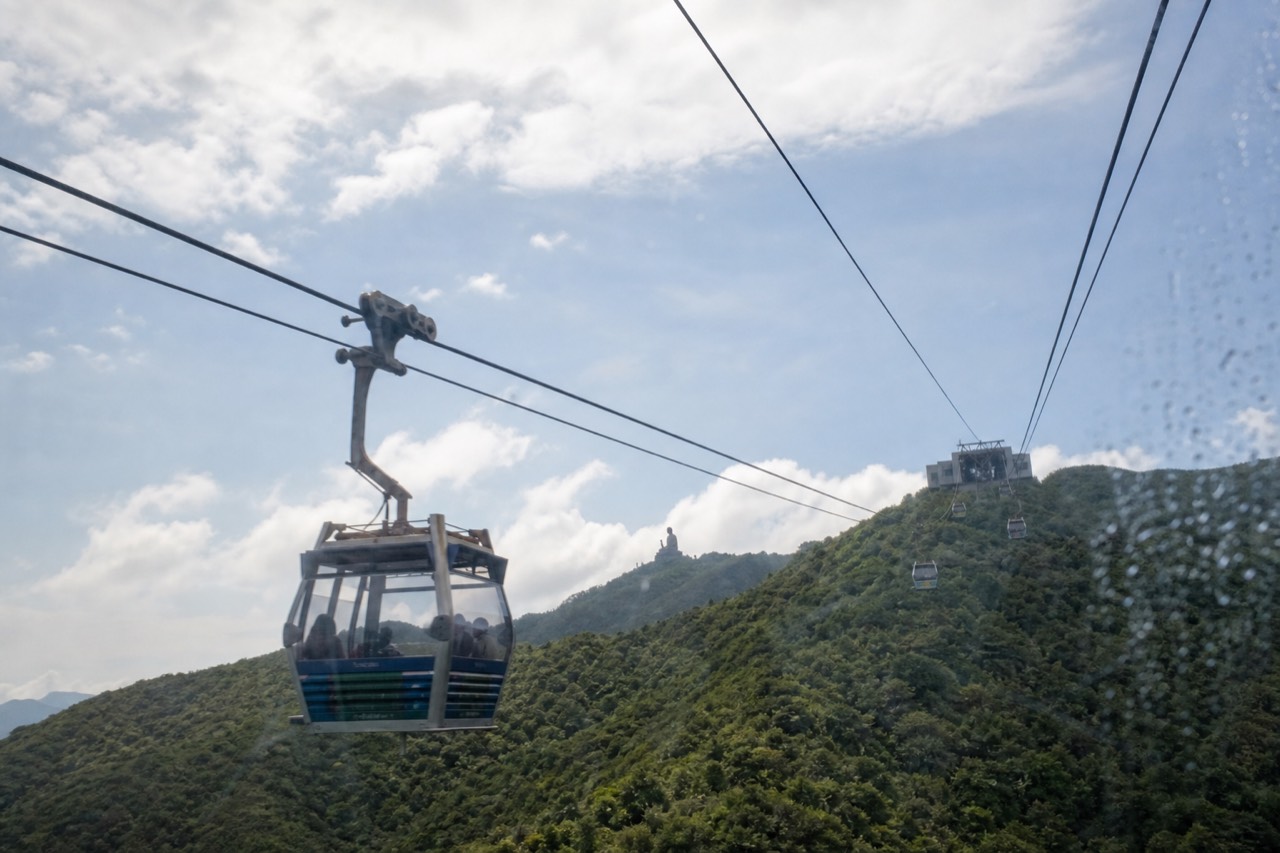 A Ngong Ping 360 cable car cabin crossing above green Lantau hills toward the upper terminal, the Tian Tan Buddha visible in the distance under a bright sky